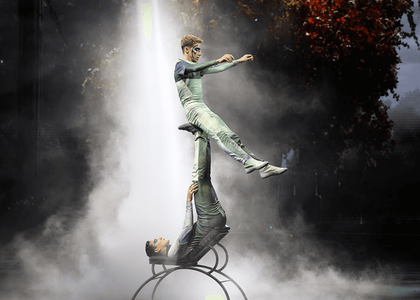 cirque artist sitting on another's feet while balancing on a wheel